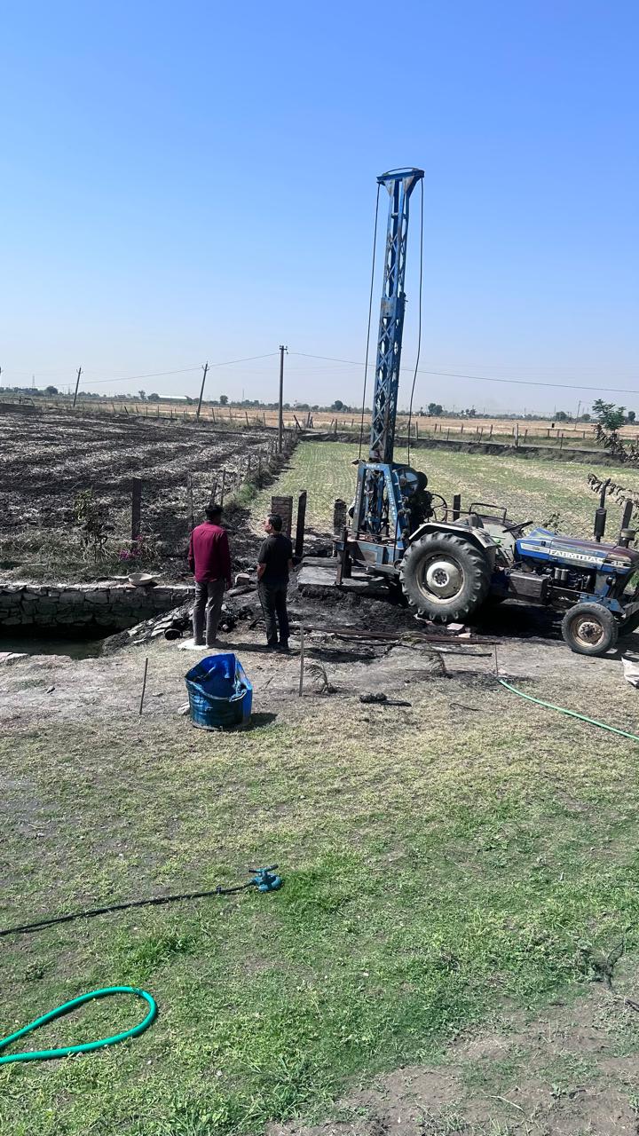 A blue drilling rig next to the tubewell, with the blackened neighbour's field on the left and the green Kota Natural Farm field on the right